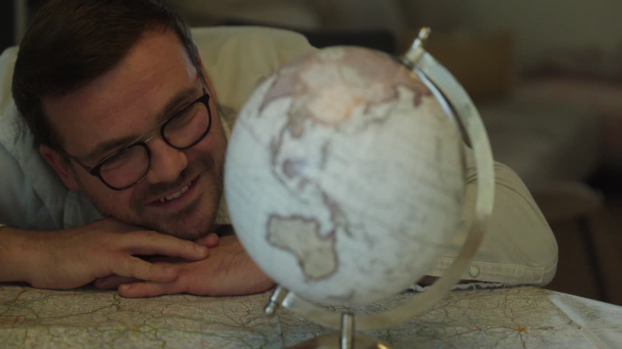 A man lies comfortably on a table, smiling as he looks at a globe, imagining destinations and feeling excitement for his future travels