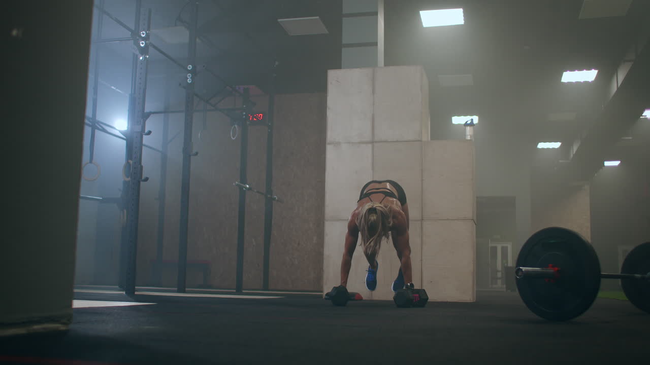 A woman performs push-UPS with dumbbells. burpee with weight. The jerk of the dumbbells over your head. A woman trains with a heavy weight.