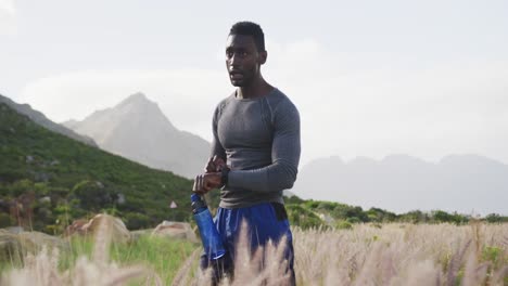 hombre afroamericano bebiendo agua mientras hace ejercicio en el campo comprobando su reloj