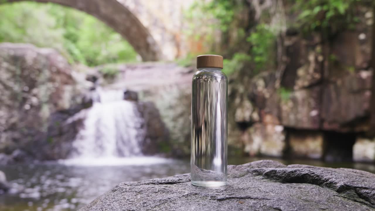 Pan out, A clear glass bottle with tan lid sits on top of a wet rock with waterfall and rock bridge in background.