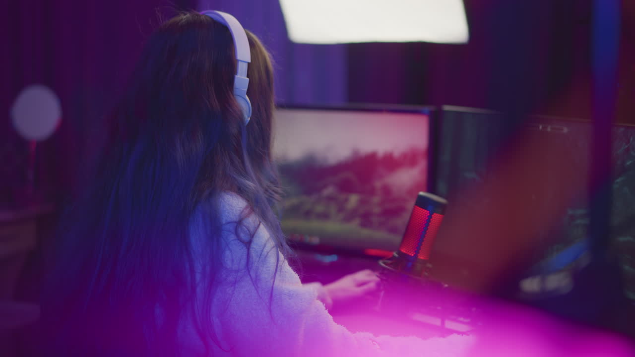 back view of woman with long cascading hair wearing white headset and fluffy sweater intensely playing video game on dual monitors with red microphone and rgb lighting in modern dark streaming studio