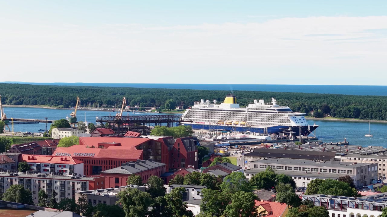 Video drone mostra il porto di Klaipėda con una grande nave da crociera, un lungomare vivace, tetti colorati e alberi rigogliosi sotto cieli sereni. La luce diurna intensa esalta le texture della città, del porto e del litorale