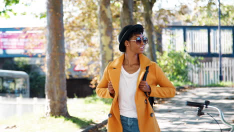 fashionable young black woman wearing a hat, sunglasses, blue jeans and a yellow pea coat walking along a treelined city street on a sunny day, looking around, smiling, front view