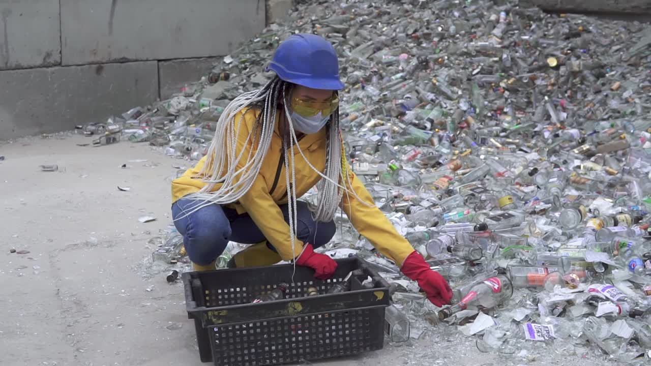 mujer con sombrero duro recogiendo botellas de vidrio blanco no rotas de la pila de vidrio roto, botellas usadas junto a la pared. niña con chaqueta amarilla en cuclillas reuniendo botellas de vidrio viejas para su reciclaje. cámara lenta
