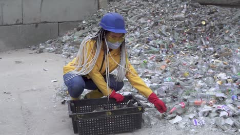 Mujer-Con-Casco-Recogiendo-Botellas-De-Vidrio-Blanco-No-Rotas-De-La-Pila-De-Vidrios-Rotos,-Botellas-Usadas-Al-Lado-De-La-Pared.-Chica-Con-Chaqueta-Amarilla-En-Cuclillas-Recogiendo-Viejas-Botellas-De-Vidrio-Para-Su-Posterior-Reciclaje.-Camara-Lenta