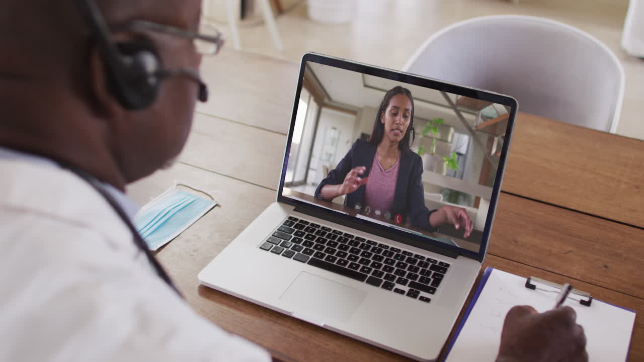 Afroamerikansk forretningsmand sidder ved skrivebord, bruger bærbar computer og har videoopkald med en kvindelig kollega.
