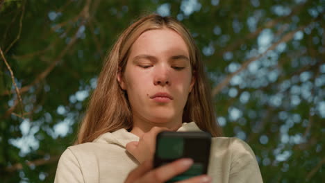 young woman in casual hoodie focuses intently on her phone while holding her hood collar, face bathed in sunlight filtering through leaves, background blurred with lush greenery