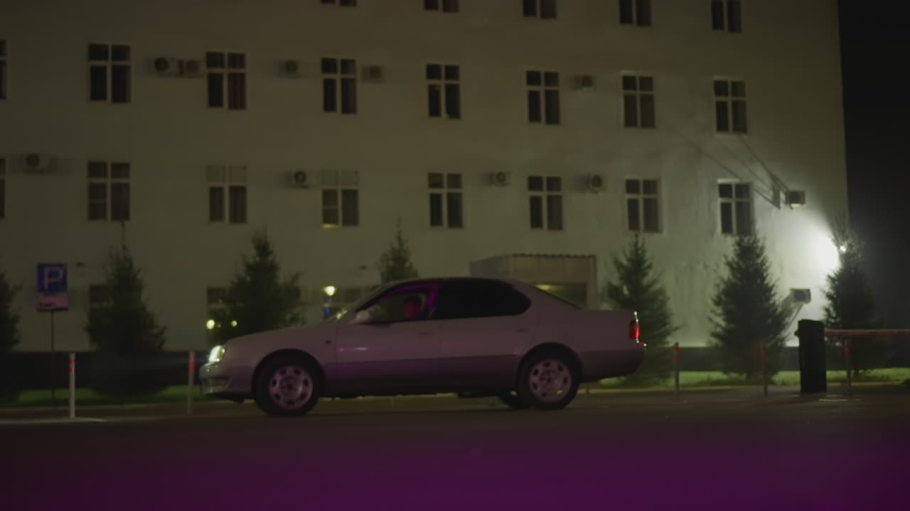 세단이 움직이는 밤의 도시 거리, 불이 켜진 대로를 지나 옆으로 운전, 아스팔트에 비치는 보라색 반사, 건물 외관, 주차 표지판, 심야 배달 풍경, 잔잔한 영화 같은 드라이브