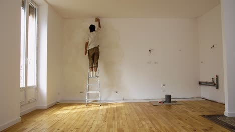 Hombre-Subiendo-Una-Escalera-De-Acero-Mientras-Pinta-Una-Habitación-Vacía-Con-Pintura-De-Cal