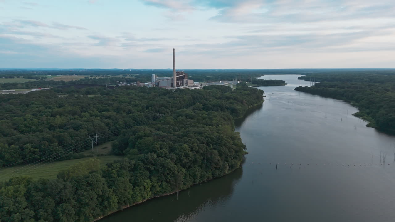Drone oversigt over kraftværk ved bred sømunding, der snor sig gennem skovklædte bakker med roligt vand under en grå himmel