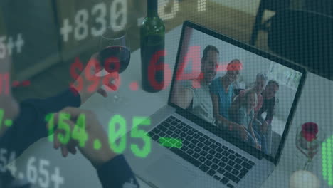 man calling family on laptop at home, holding wine glass with floating green red finance data
