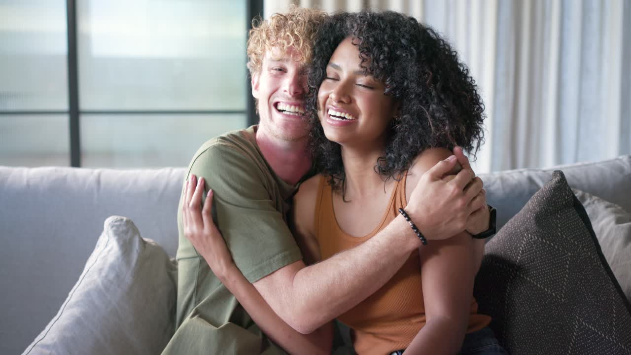 Couple sitting on gray sofa, man reaching for woman's knee, woman adjusting hair, posing for camera