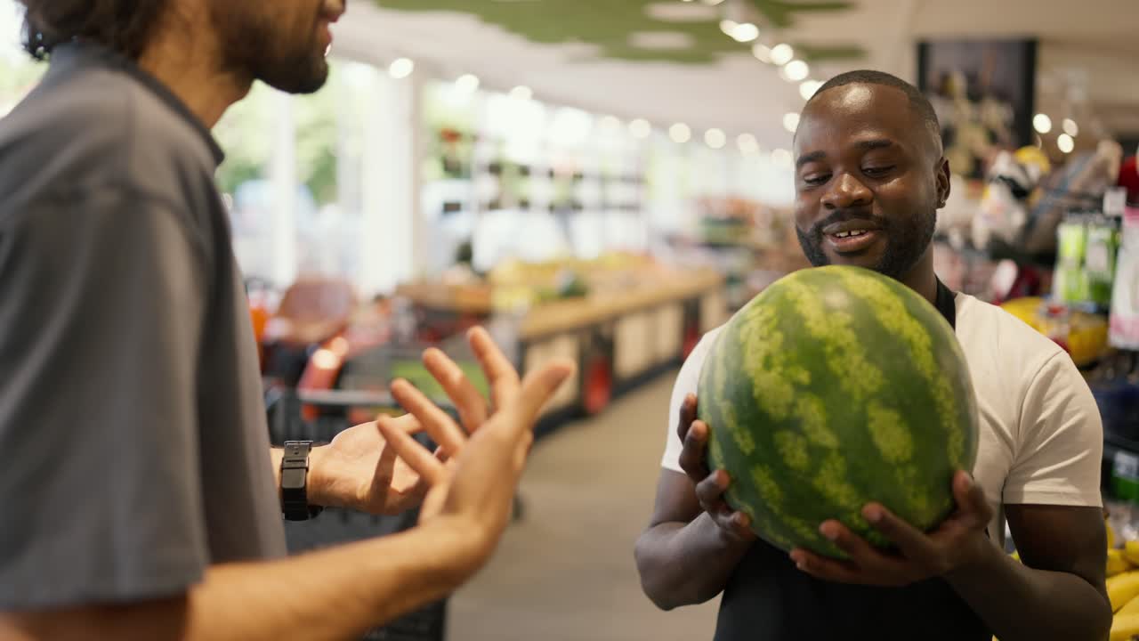 a man with Black skin in a black apron holds a watermelon in his hands and talks about it. A store customer asks a consultant about watermelon