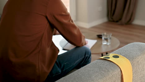 back view of an unrecognizable blind man sitting on the sofa and reading a braille book