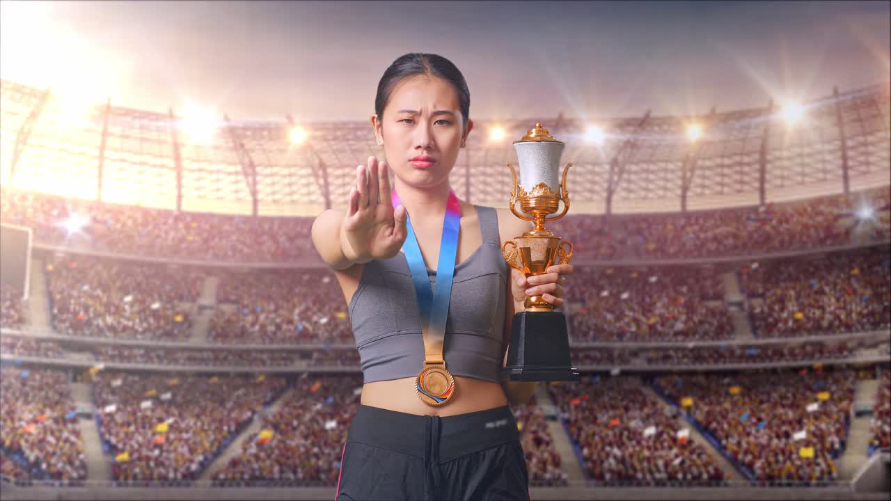 mujer asiática con una medalla de oro y trofeo desaprueba sin señal de mano y sonriendo a la cámara en el estadio