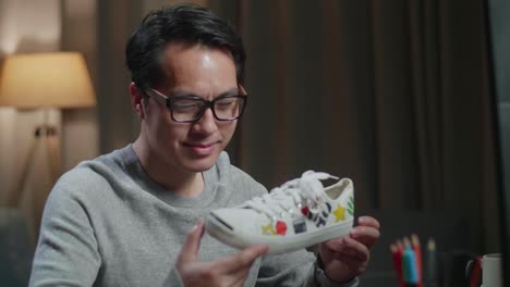 primer plano de un sonriente diseñador de calzado masculino asiático sosteniendo una zapatilla de deporte de patrones coloridos y mostrando el pulgar hacia arriba en el gesto de la cámara mientras trabaja en una computadora de escritorio en casa