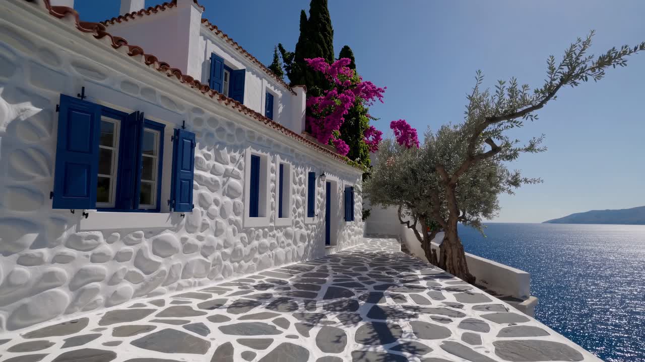 Whitewashed stone house with blue shutters and tiled terrace overlooking the Aegean Sea, featuring vibrant bougainvillea and an olive tree, bathed in sunlight