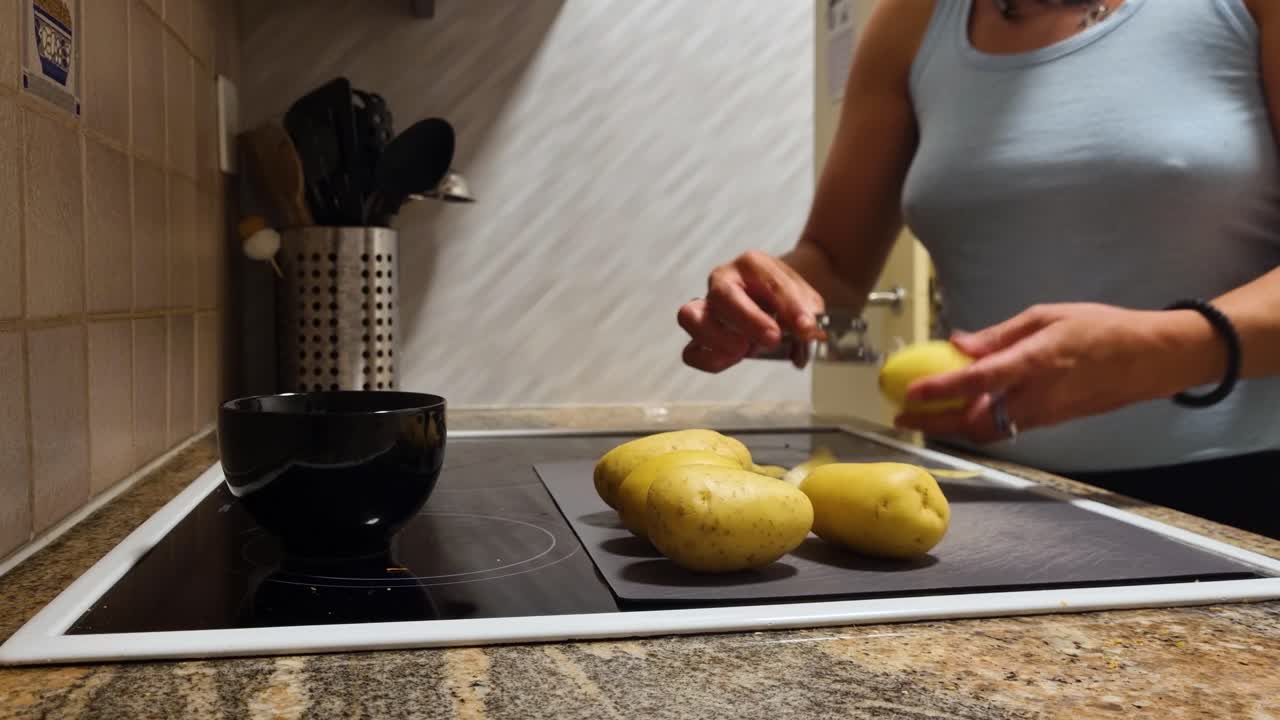 Close-up of a woman in a blue tank top peeling potatoes in her home kitchen in Bern, Switzerland, preparing ingredients for a healthy homemade meal