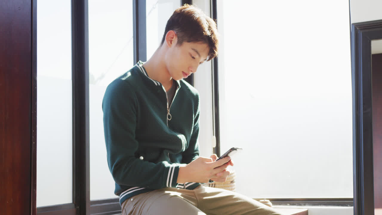집의 창문 근처에 앉아 스마트폰을 사용하는 아시아 소년