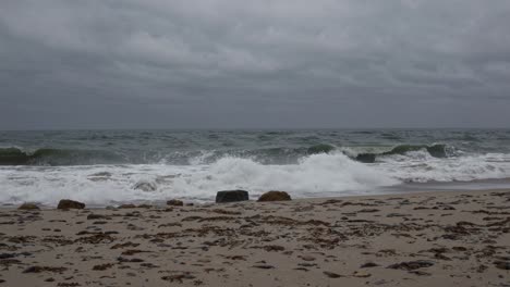 ocean waves rolling powerfully across sandy shoreline, revealing abandoned tire resting amid foam and cloudy gray sky, depicting environmental landscape and natural seascape dynamics