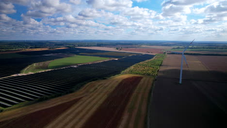 Aerial-flight-towards-rotating-wind-turbine-and-solar-panel-farm-on-countryside-fields-at-cloudy-day