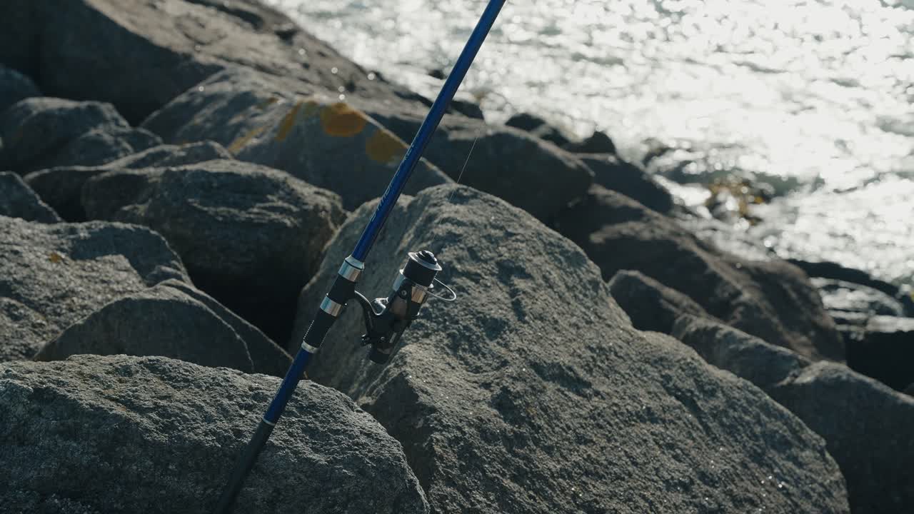 Close up of a blue fishing rod secured among coastal rocks by shimmering water