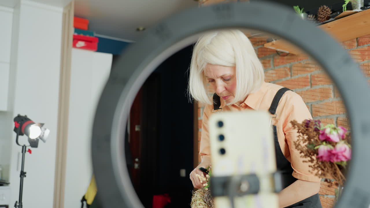 Female florist with blonde hair wearing apron arranging dried flowers while being filmed through smartphone on tripod with ring light in creative studio setup professional lighting equipment