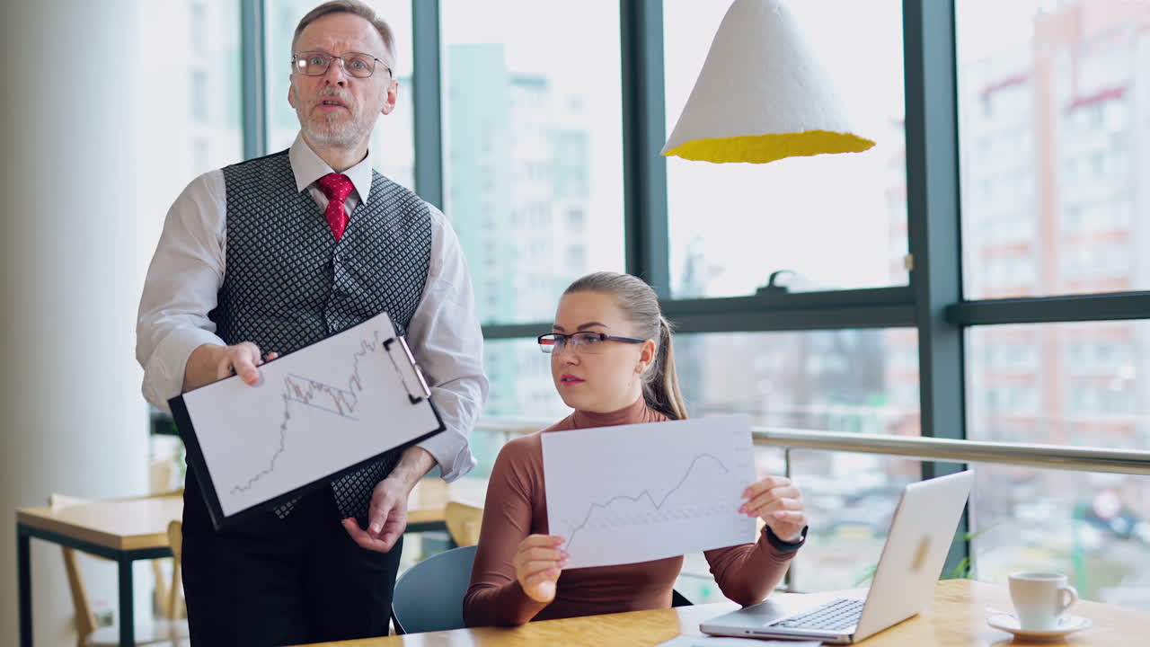 Elderly businessman and a secretary in the office. Attractive company worker sitting at the desk and talking to her boss about some graphics.
