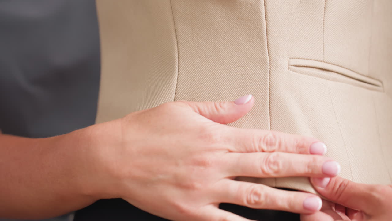 Side view beige waistcoat on black mannequin while seamstress fingers smooth seam line, adjusting fit to perfection, inspecting fabric grain and dart placement during focused tailoring session studio