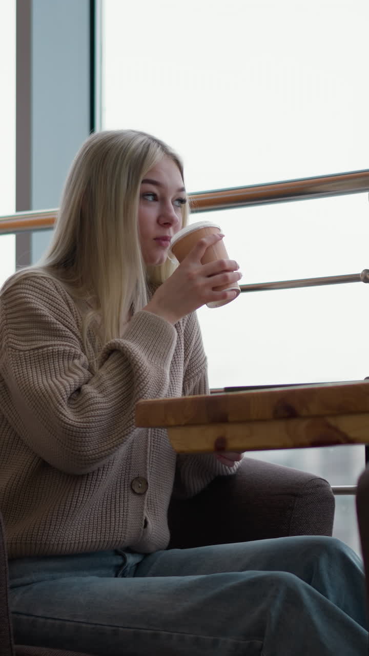 멋진 여성이 커피를 마시며, 장식 된 목조 테이블과 유리 창을 배경으로 현대적인 쇼핑몰 카페에 앉아 있으며, 부드러운 조명과 도시 풍경을 가진 조용한 분위기입니다.