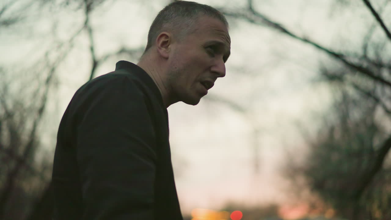 A man in a green jacket smokes a cigarette in a dimly lit forest at dusk, the scene captures the solitary moment as smoke escapes from his mouth and nose
