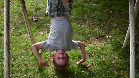 young boy hangs upside down smiling outdoors with background of leaves and grass