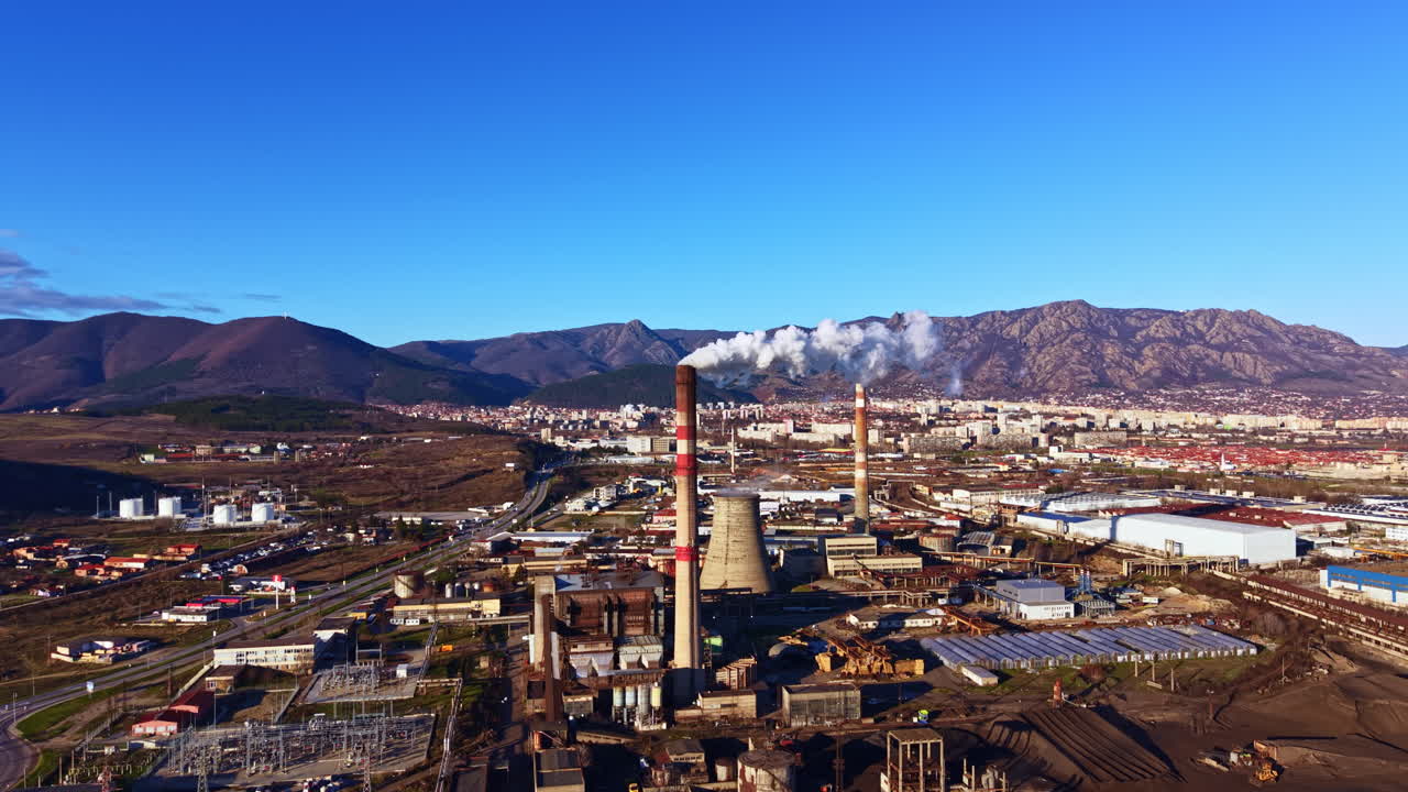 Fumaça sobe de torres industriais em uma cidade cercada por montanhas durante o dia claro