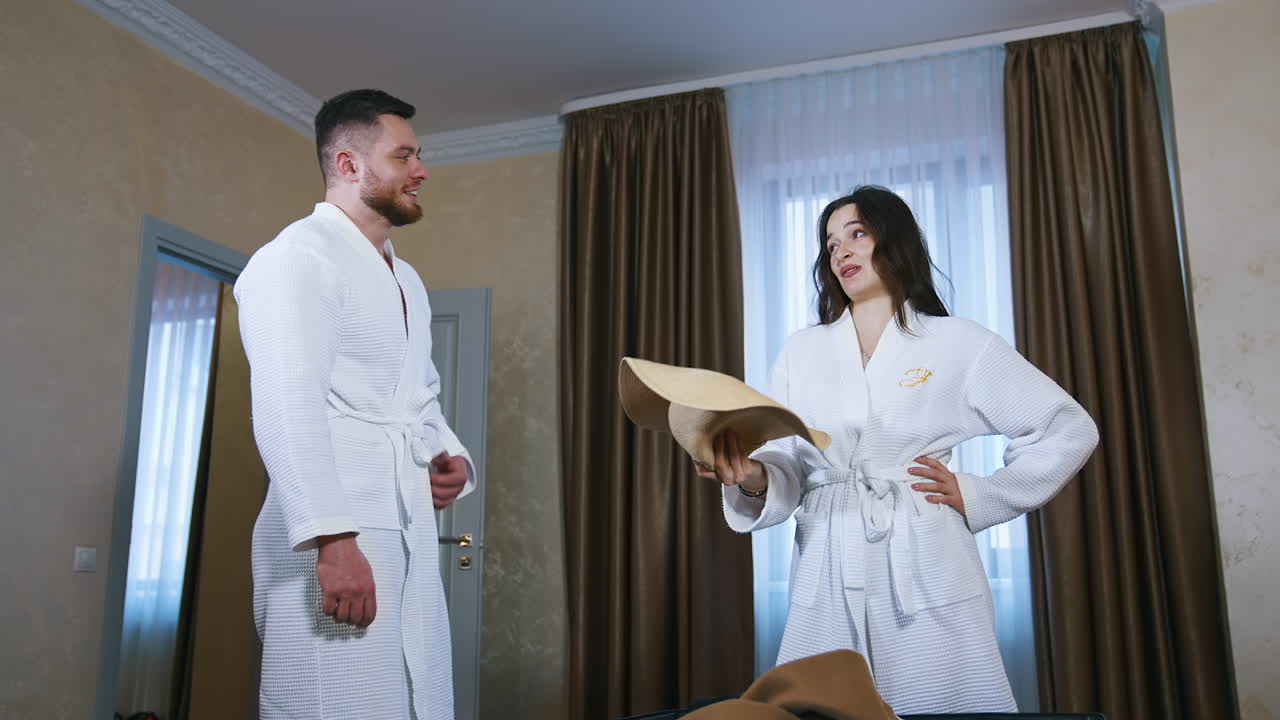 Cheerful couple in the hotel. Young man and woman in white dressing gowns in the apartment. Tired woman waving her hat and talking to her husband.