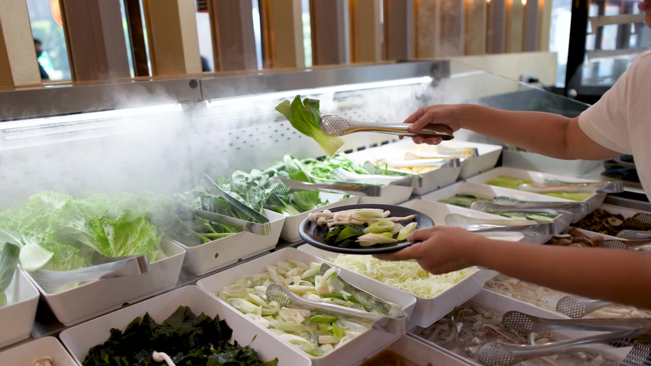 Una persona usa le pinze per prendere verdure fresche da un espositore a buffet refrigerato e fumante.