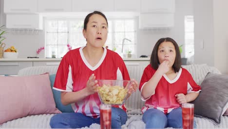Video-Einer-Glücklichen-Asiatischen-Mutter-Und-Tochter,-Die-Chips-Isst-Und-Sich-Ein-Spiel-Im-Fernsehen-Ansieht