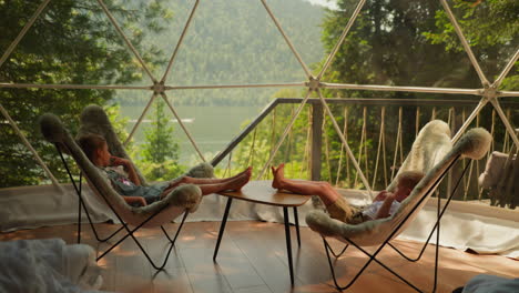 niños en sillas suaves bajo la cúpula de glamping. niño y niña descalzos se sientan en sillones esponjosos cerca de la ventana panorámica. niños pequeños descansan en el campamento en vacaciones