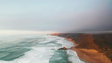 Portugal beach cliffs on Atlantic ocean at sunset from high aerial drone forwards shot with rough waves near rocky outcrop in water