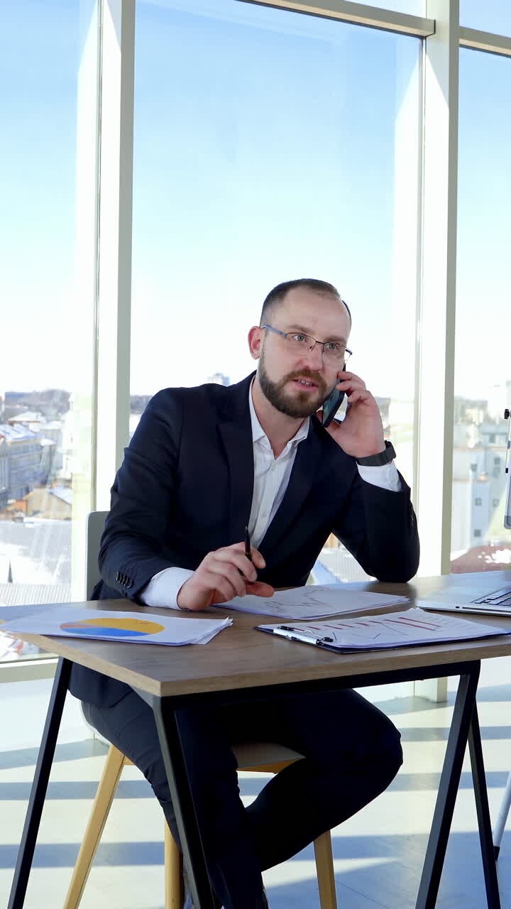 Professional entrepreneur working at office centre. Businessman in suit sitting at his workplace and talking the phone on large windows background. Vertical video