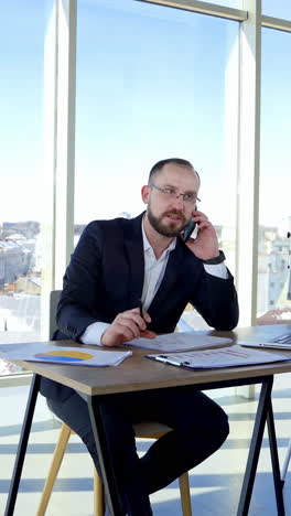 professional entrepreneur working at office centre. businessman in suit sitting at his workplace and talking the phone on large windows background. vertical video