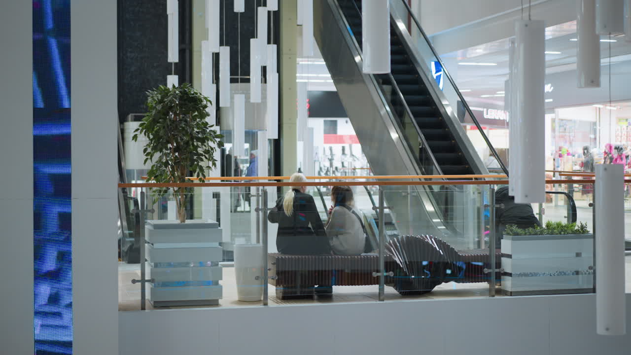 Two girls seated across on bench in modern shopping mall near escalator with decorative lighting columns potted plant and architectural details showing casual urban lifestyle