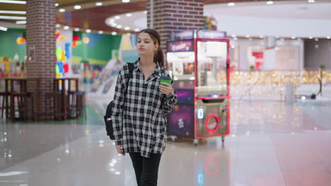 young girl in casual plaid shirt walks through shopping mall while sipping juice, she carries backpack, looks around curiously, and enjoys relaxed moment in vibrant indoor shopping environment