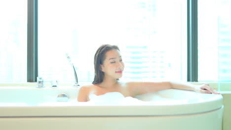 편안하게 목욕하는 아시아 여성이 유리벽이 있는 고급 아파트 단지에서 거품이 가득한 욕조에 누워 있다
