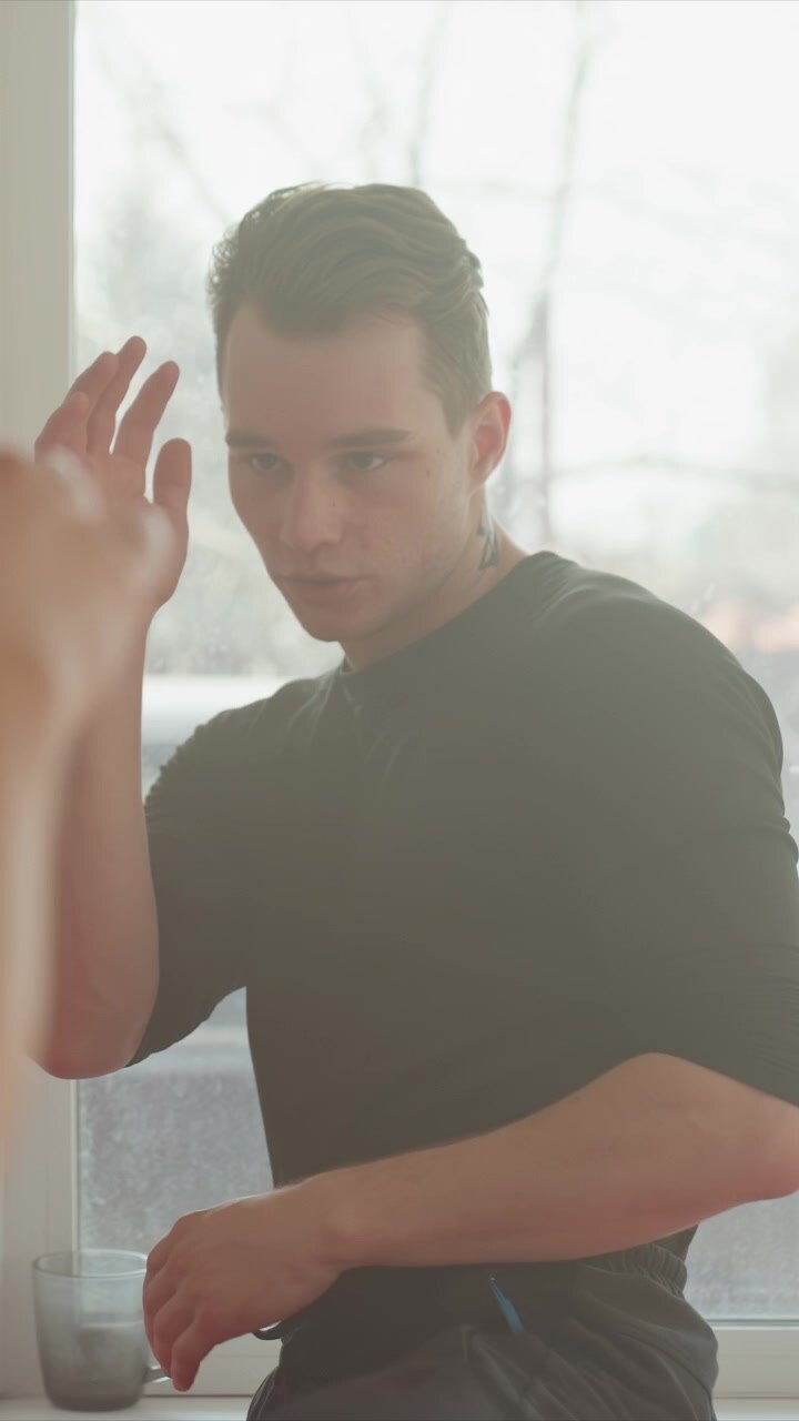 Young man with short brown hair and black shirt makes. Tattoo visible at back of neck near ear with earring. Scene begins mostly obscured by darkness then gradually shows