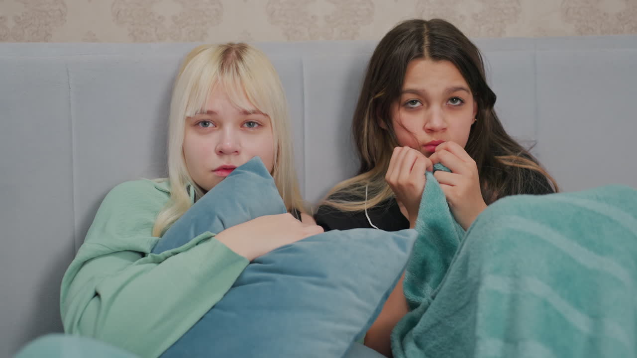 young girls sitting close together on bed watching horror movie, looking frightened with anxious facial expressions, one clutching pillow while other bites blanket