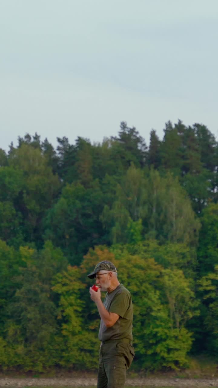 Vertical video of senior men taking a bite into a red apple. Eating outdoors with verdant forest in the background.