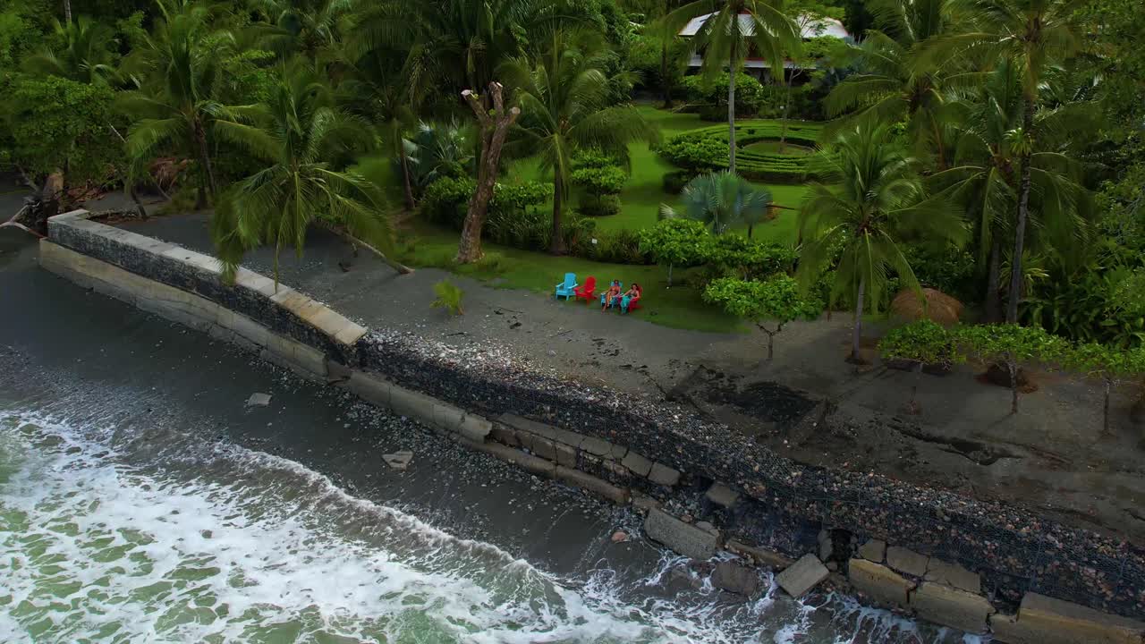 Drone pulls back from two women at a small beachfront resort in Costa Rica. Ocean waves crash nearby