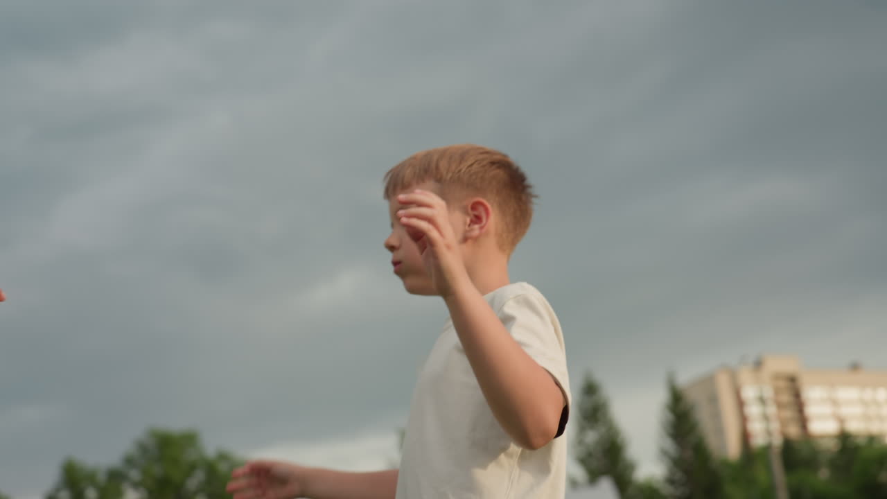 sky view of boy running toward mom in park while chewing snack, eyes closed in playful moment, summer grass and gentle breeze in background, candid family interaction showing energy and affection