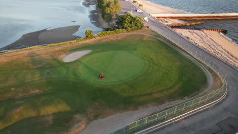 an aerial view of a person riding a lawnmower cutting the grass of a golf course between a beach and a pond during sunrise