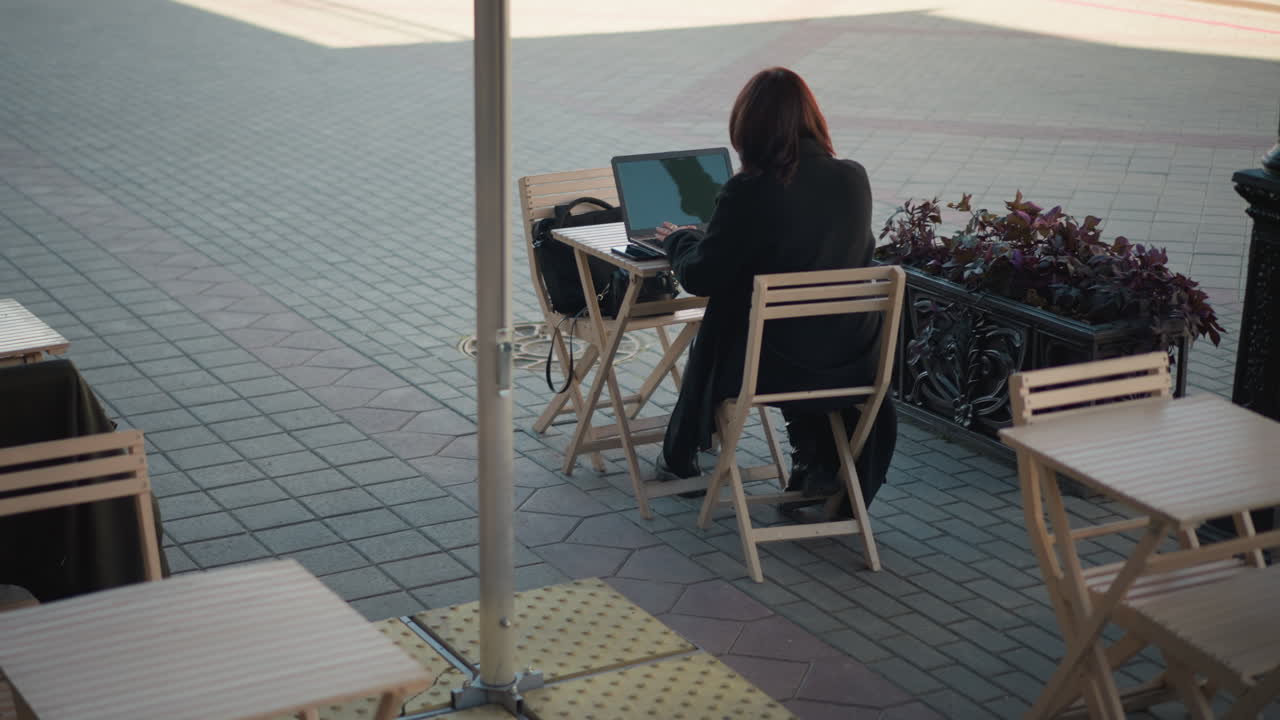 mujer profesional escribiendo en una computadora portátil al aire libre, con el reflejo del poste visible en la pantalla, trabajando en un entorno urbano en una cafetería rodeada de sillas de madera, plantas en macetas y una pasarela pavimentada